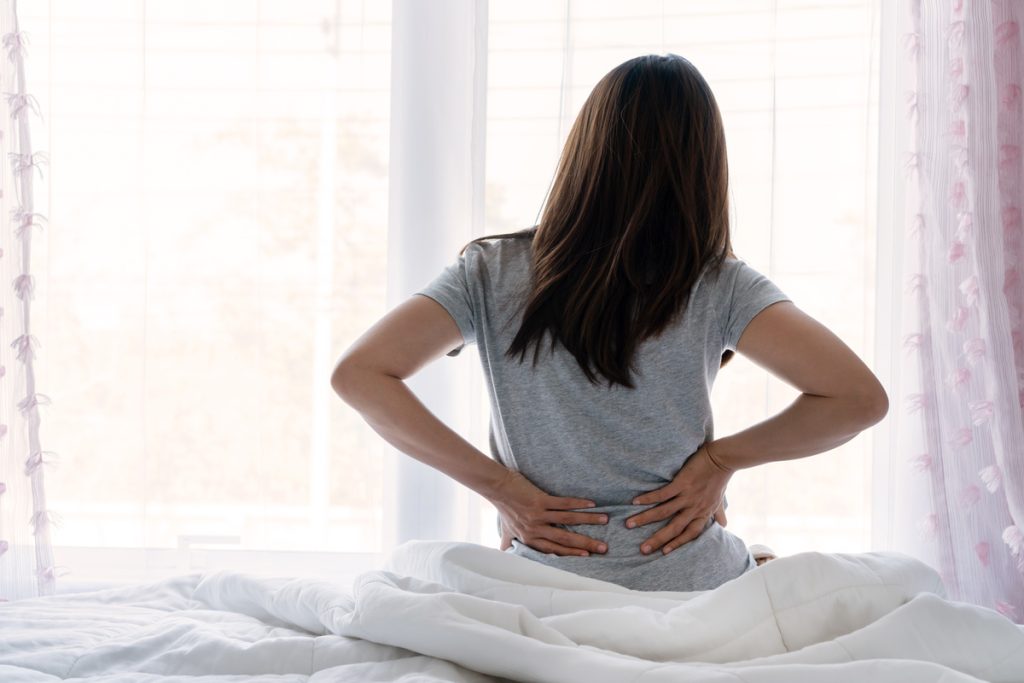 Triste joven asiática tocando espalda sintiendo dolor de espalda malestar matutino dolor lumbar bajo riñón muscular sentarse en la cama después de mal sueño despertar en colchón incómodo flexión. Concepto de mujer estirándose sufriendo dolor de espalda repentino.