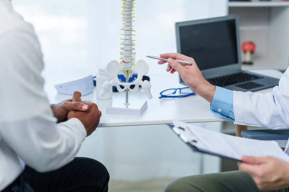 Primer plano de un médico y un paciente en un consultorio médico, modelo de columna vertebral, posiblemente hablando de una enfermedad o síndrome de la columna vertebral con el paciente.